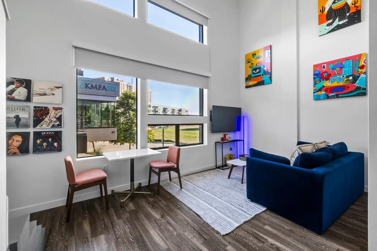 Sala de estar moderna con ventanas grandes, un sofá azul, una mesa de comedor pequeña con sillas rosas, obras de arte y una televisión.