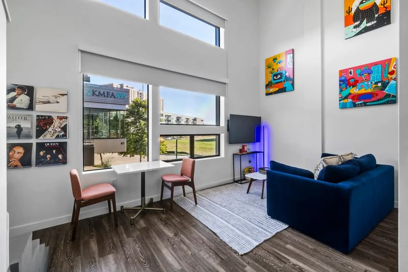 Modern living room with large windows, a blue sofa, a small dining table with pink chairs, artwork, and a TV.