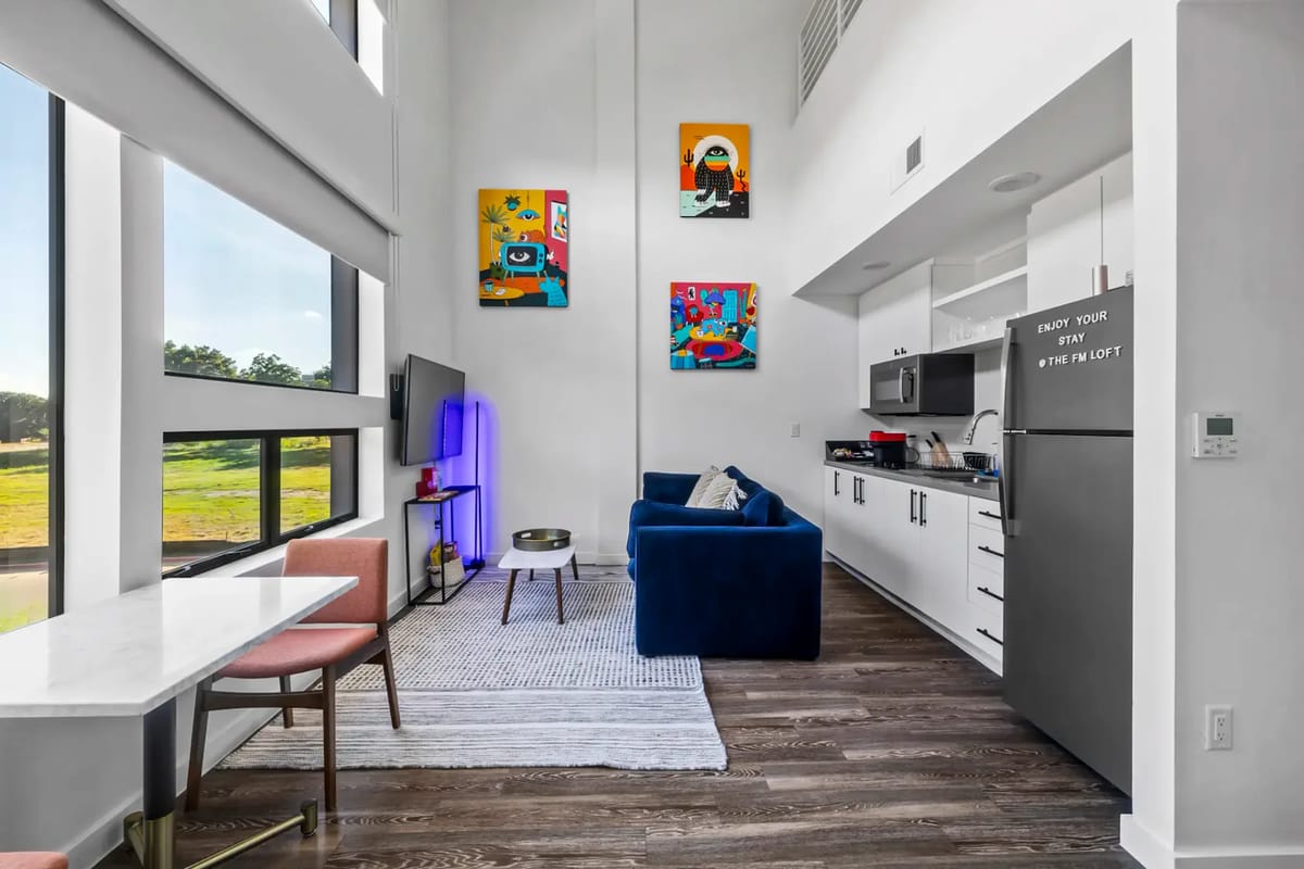 Sala de estar moderna con ventanas grandes, arte de colores vivos en las paredes, un sofá azul y un área de cocina.