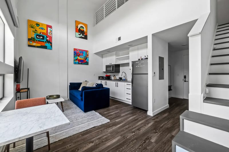 Modern apartment interior with a blue sofa, kitchen area, and staircase. Colorful art decorates white walls and wood flooring runs throughout.