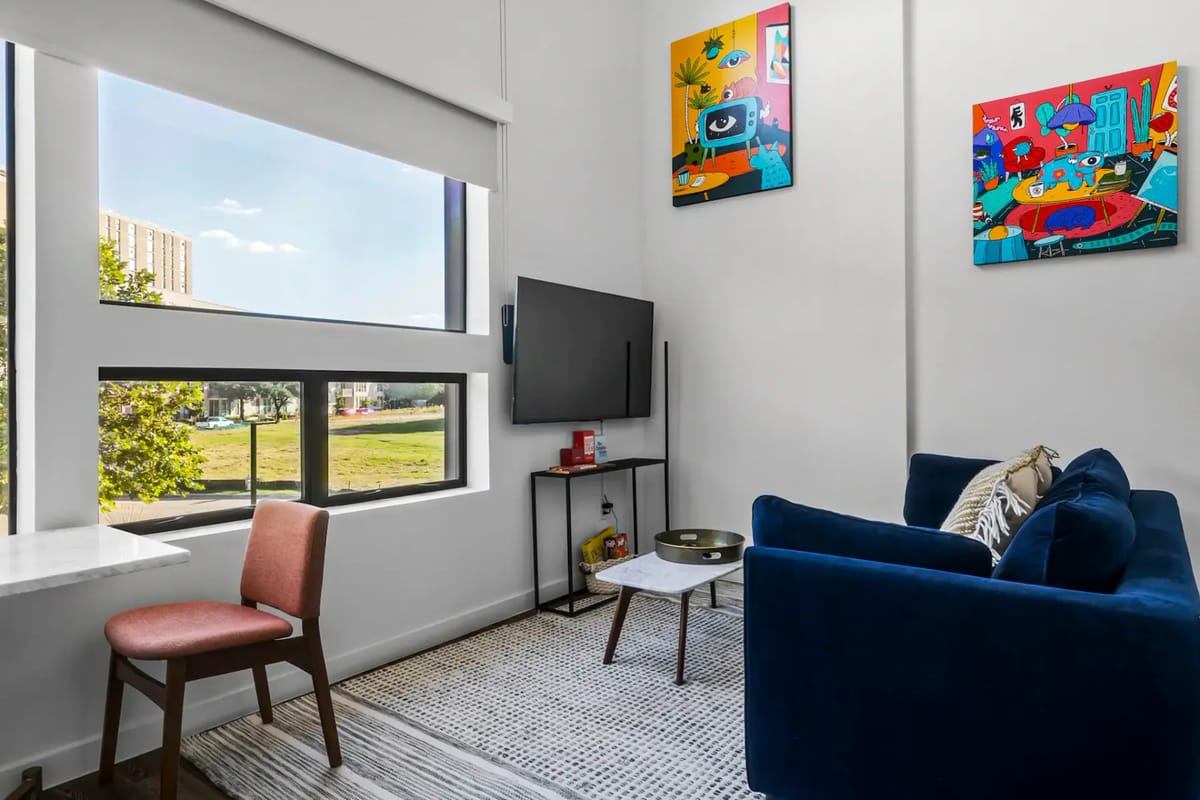 Sala de estar moderna con un sofá azul, televisión montada en la pared, obras de arte coloridas, ventanas grandes, una mesa pequeña y una silla junto a un escritorio.