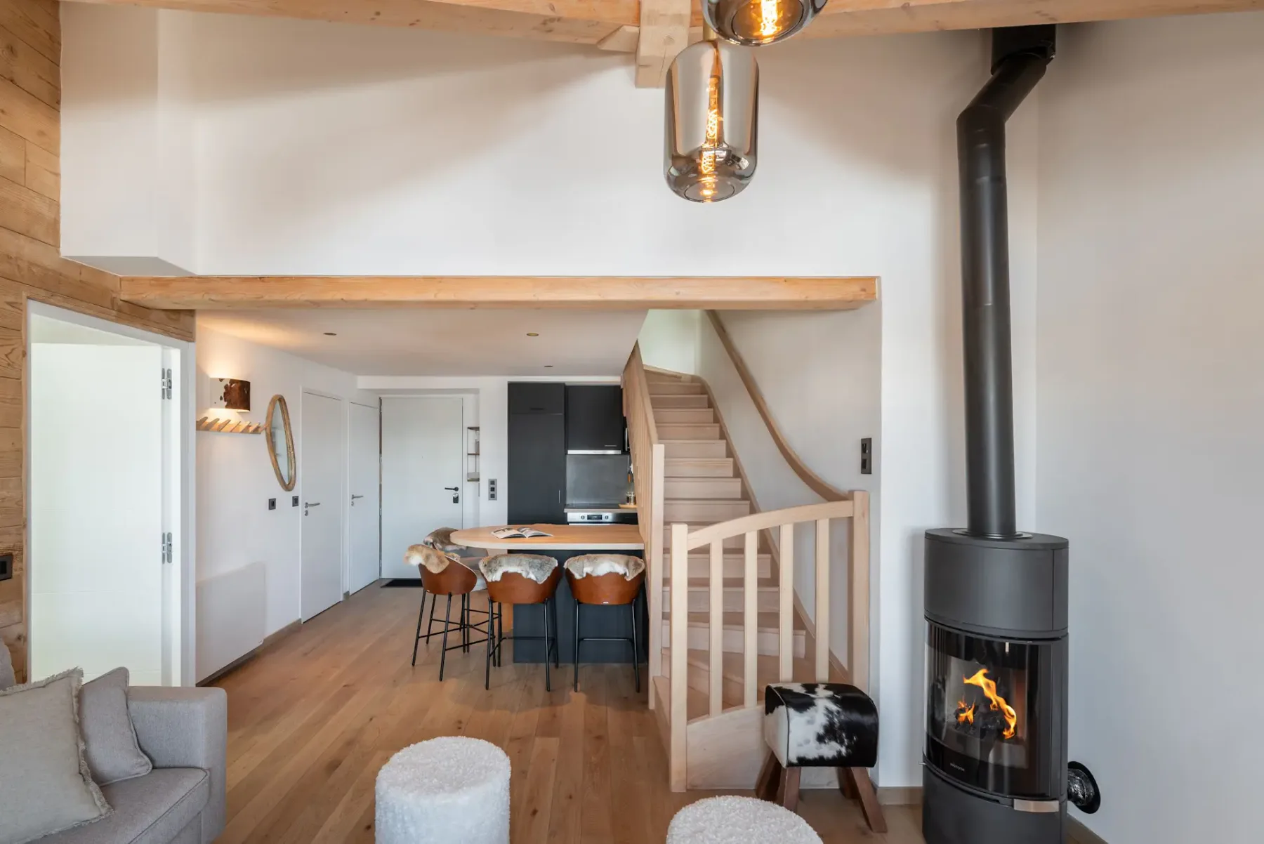 Salón moderno con vigas de madera, una ventana grande y vistas a la montaña. Cuenta con un sofá, dos sillones, una mesa de centro y una alfombra estampada. Una escalera conduce a un nivel superior.