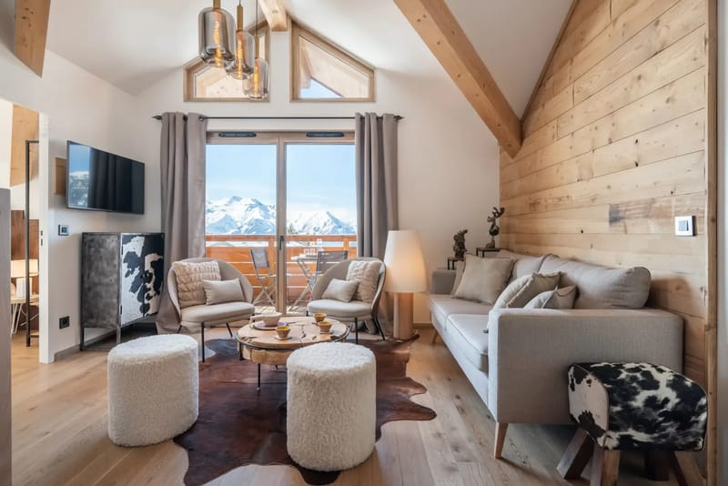Modern living room with wooden beams, large window, and mountain view. Features a sofa, two armchairs, a coffee table, and a patterned rug. A staircase leads to an upper level.