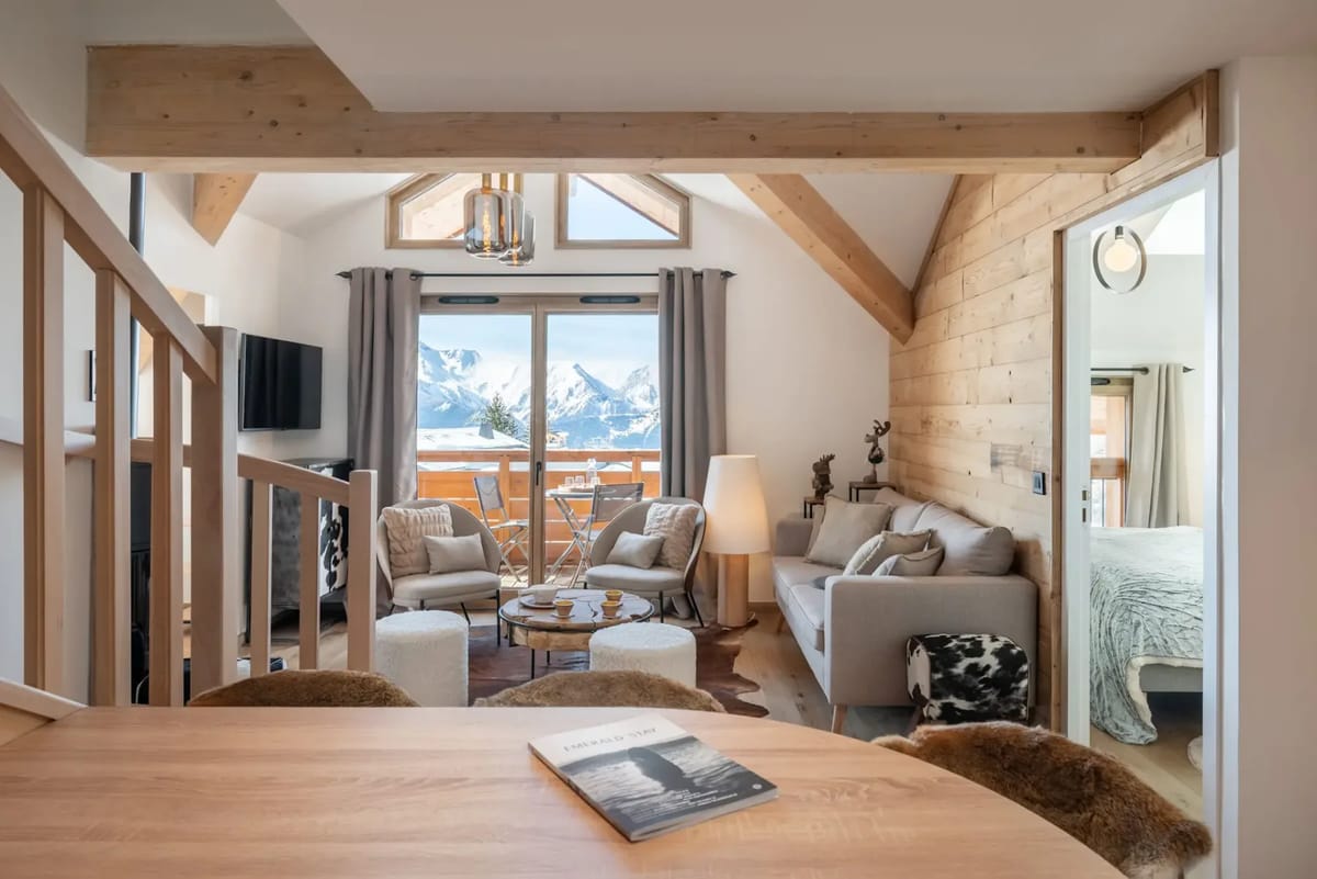 Salón moderno con vigas de madera, una ventana grande y vistas a la montaña. Cuenta con un sofá, dos sillones, una mesa de centro y una alfombra estampada. Una escalera conduce a un nivel superior.