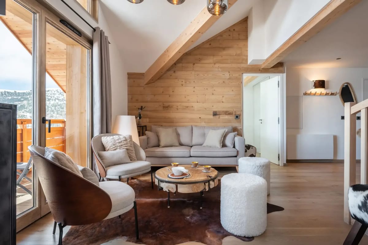 Salón moderno con vigas de madera, una ventana grande y vistas a la montaña. Cuenta con un sofá, dos sillones, una mesa de centro y una alfombra estampada. Una escalera conduce a un nivel superior.