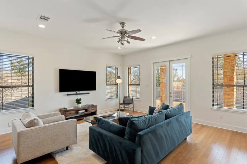 Bright living room with a blue sofa, beige armchair, TV, ceiling fan, large windows, and wooden floor. Door leads to an outdoor patio.