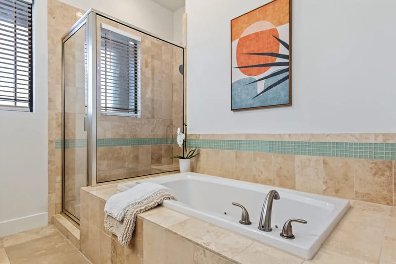 Modern bathroom with a tiled bathtub, nearby shower, decorative wall art, and small plant. A towel is draped over the tub edge.