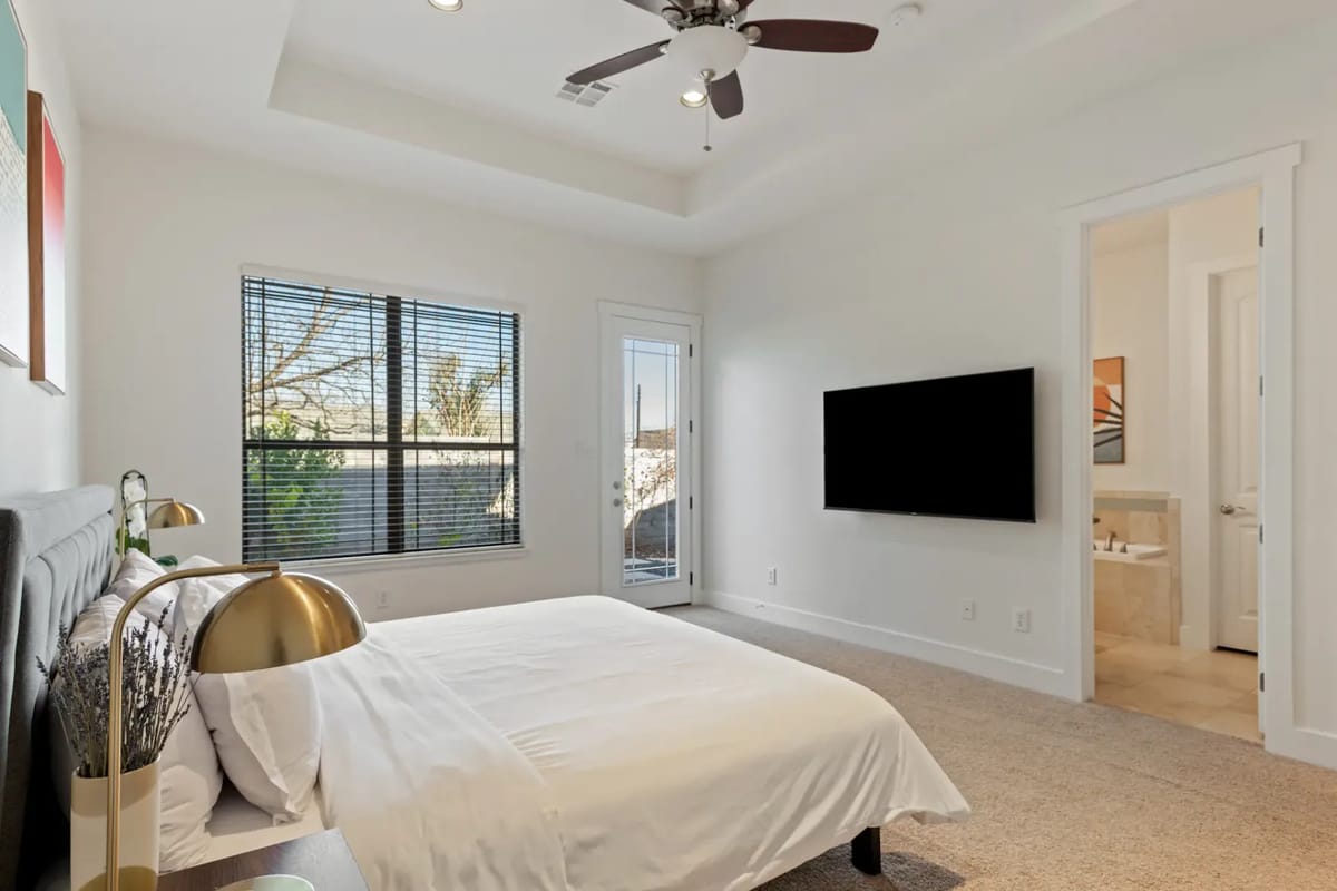 Dormitorio moderno con una cama blanca, dos lámparas doradas, un televisor montado en la pared y ventanas grandes con persianas. Un vano abierto conduce a un baño.