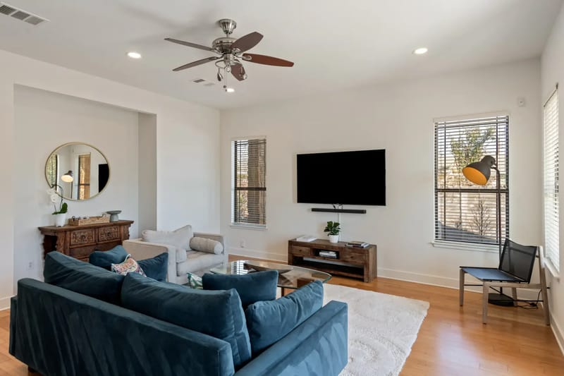 Bright living room with a blue sofa, beige armchair, TV, ceiling fan, large windows, and wooden floor. Door leads to an outdoor patio.
