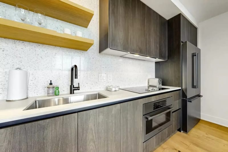Cocina moderna con gabinetes de madera, fregadero de acero inoxidable, estufa eléctrica, horno, tostadora y revestimiento de mármol contra salpicaduras.