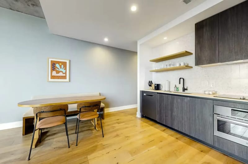 Sala de estar moderna con pisos de madera, una mesa de comedor, sillas, un sofá, ventanas grandes y obras de arte en las paredes.