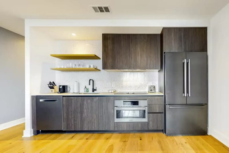 Cocina moderna con gabinetes de madera oscura, electrodomésticos de acero inoxidable, estantes abiertos y piso de madera clara.
