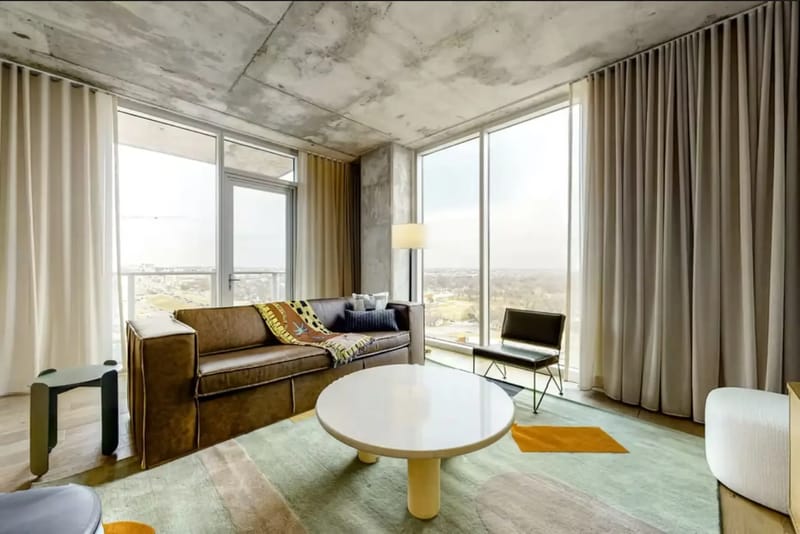 Sala de estar moderna con un sofá marrón, una mesa de centro circular, ventanas de piso a techo y vistas al paisaje urbano.
