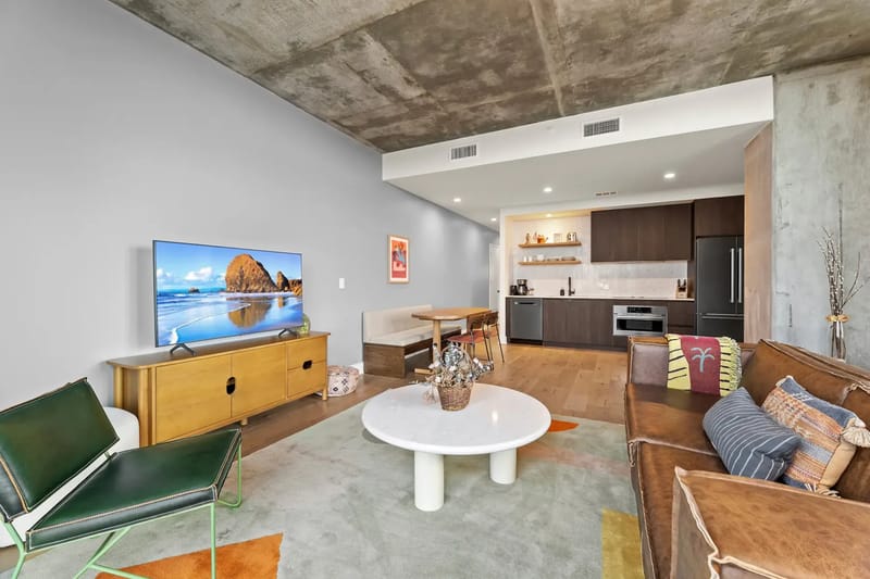 Sala de estar moderna con un sofá de cuero marrón, sillas verdes, una mesa de centro redonda blanca, un televisor montado en la pared y una cocina al fondo.