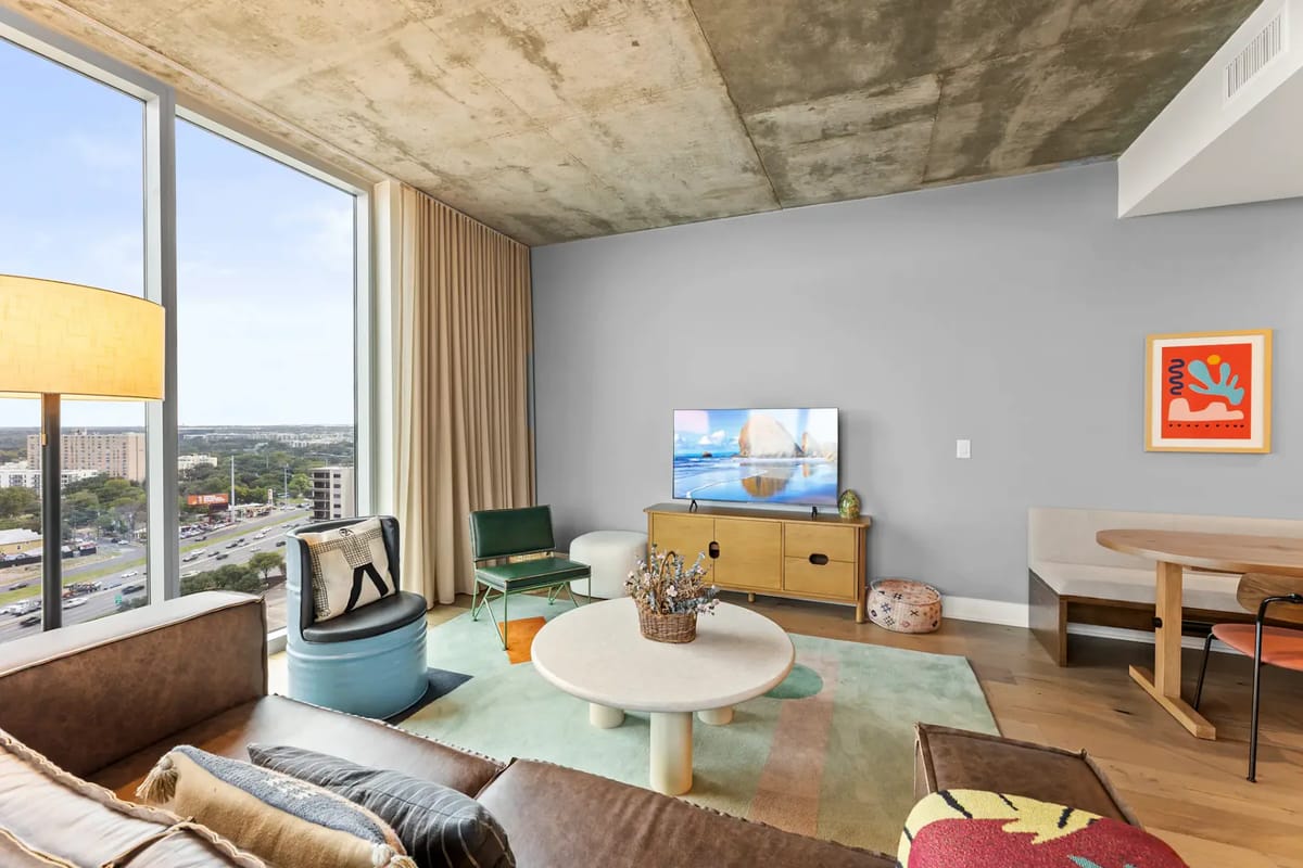 Sala de estar moderna con una ventana grande, un sofá beige, una mesa de centro redonda, un televisor, una alfombra colorida y decoración ecléctica.