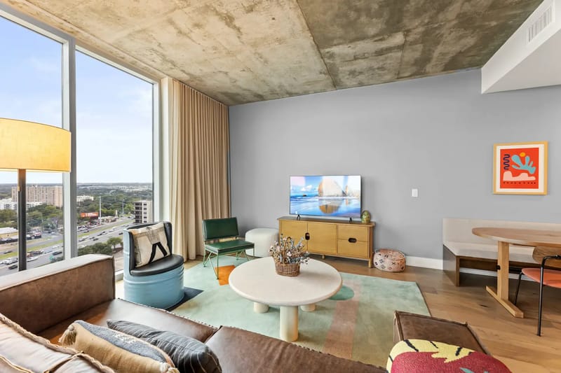 Sala de estar moderna con una ventana grande, un sofá beige, una mesa de centro redonda, un televisor, una alfombra colorida y decoración ecléctica.