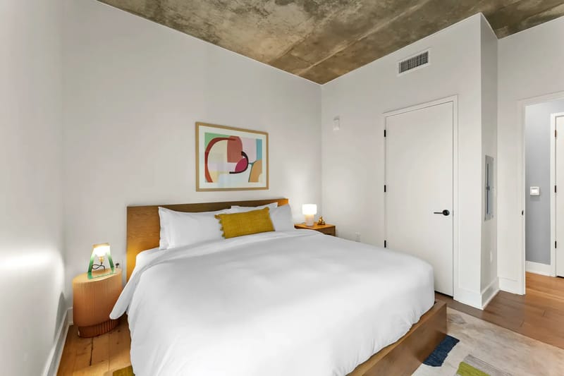 Dormitorio acogedor con una cama grande, ropa de cama blanca, mesas de noche, lámparas, arte abstracto y una alfombra.