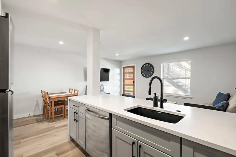 Cocina y sala de estar modernas con una isla blanca, un grifo negro, una mesa de comedor, un reloj de pared y un sofá.