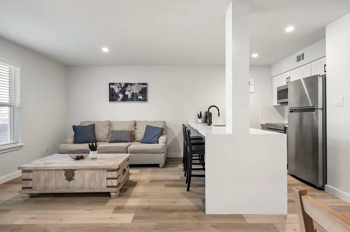 Espacio de estar moderno de planta abierta con un sofá beige, una isla de cocina, electrodomésticos de acero inoxidable y una mesa de centro de madera.