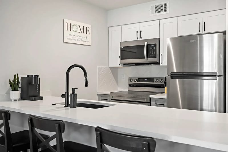Cocina moderna con electrodomésticos de acero inoxidable, gabinetes blancos, un grifo negro y una encimera blanca.
