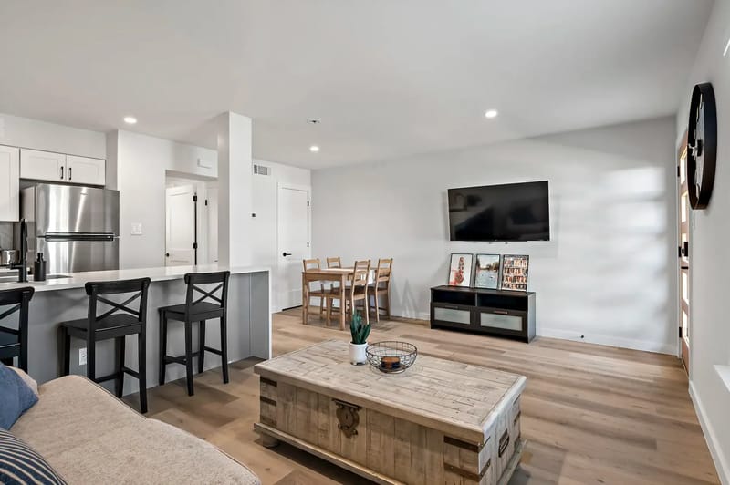 Sala de estar moderna con pisos de madera, un televisor montado en la pared, una mesa de comedor y una cocina con taburetes de barra.