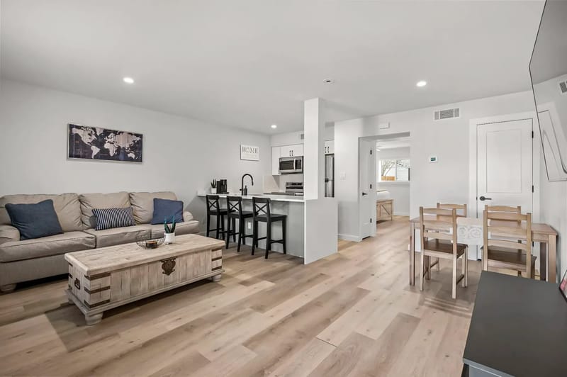 Sala de estar moderna de concepto abierto con un sofá beige, una mesa de centro de madera, taburetes junto a la barra de la cocina y una mesa de comedor sobre pisos de madera clara.