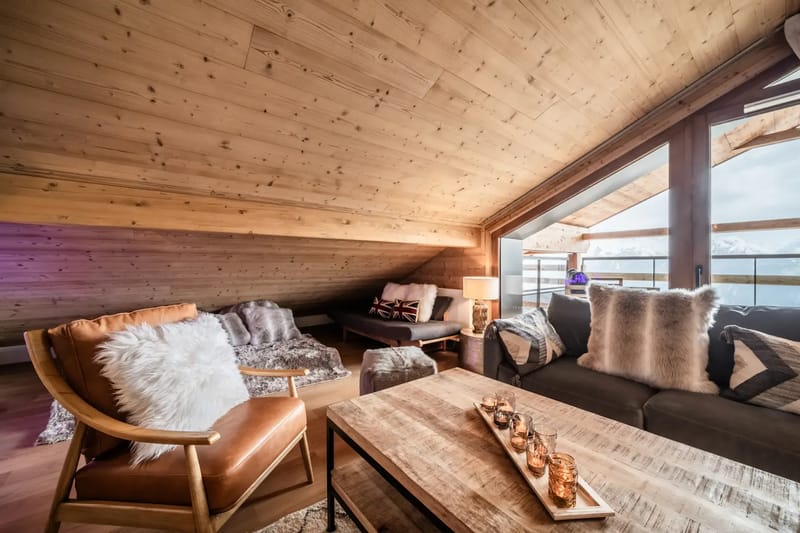 Acogedora sala de estar de una cabaña de montaña con paredes y techo de madera, sofás mullidos, un sillón y grandes ventanas con vistas a un paisaje nevado.