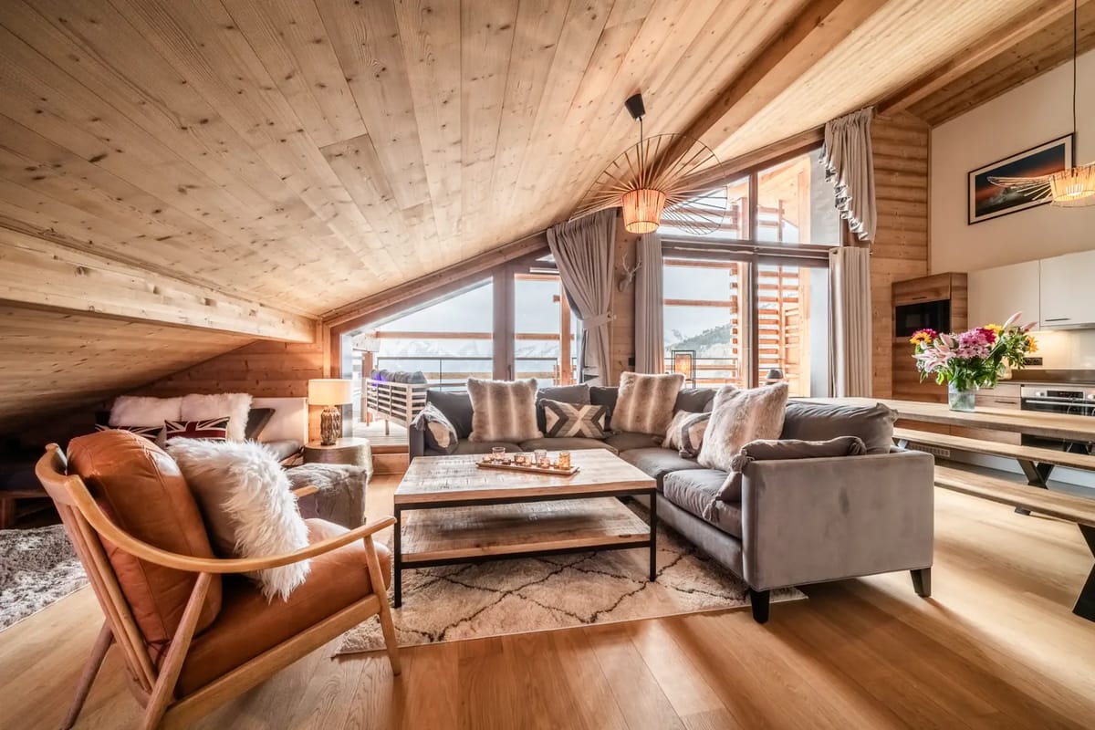 Acogedora sala de estar de una cabaña de montaña con paredes y techo de madera, sofás mullidos, un sillón y grandes ventanas con vistas a un paisaje nevado.