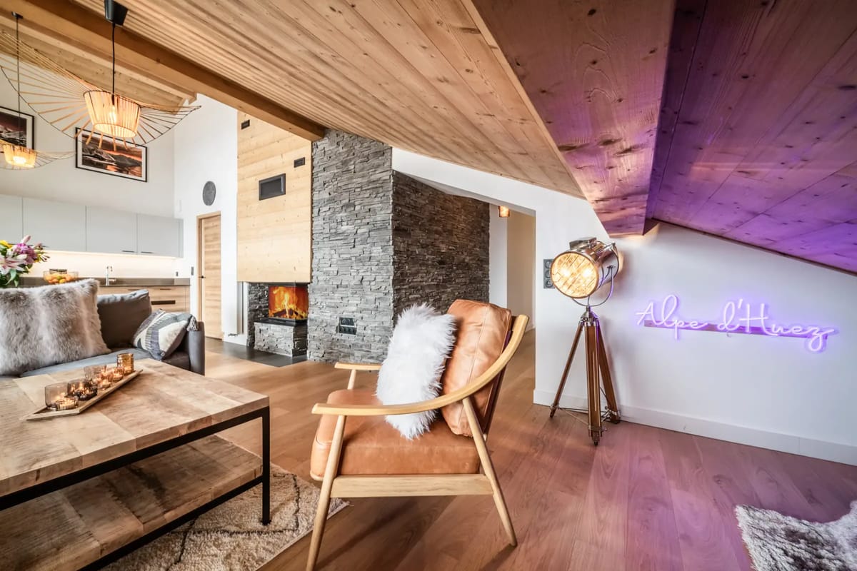 Acogedora sala de estar de una cabaña de montaña con paredes y techo de madera, sofás mullidos, un sillón y grandes ventanas con vistas a un paisaje nevado.