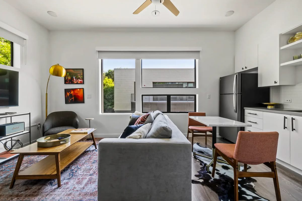 Modern living room with a gray sofa, wall-mounted TV, large windows, a wooden coffee table, and framed art.