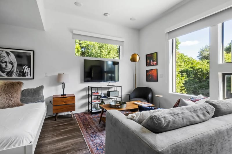 Sala de estar moderna con un sofá gris, un televisor montado en la pared, ventanas grandes, una mesa de centro de madera y obras de arte enmarcadas.