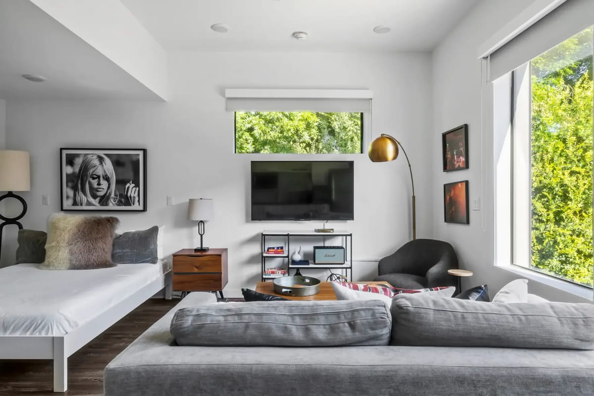 Modern living room with a gray sofa, wall-mounted TV, large windows, a wooden coffee table, and framed art.
