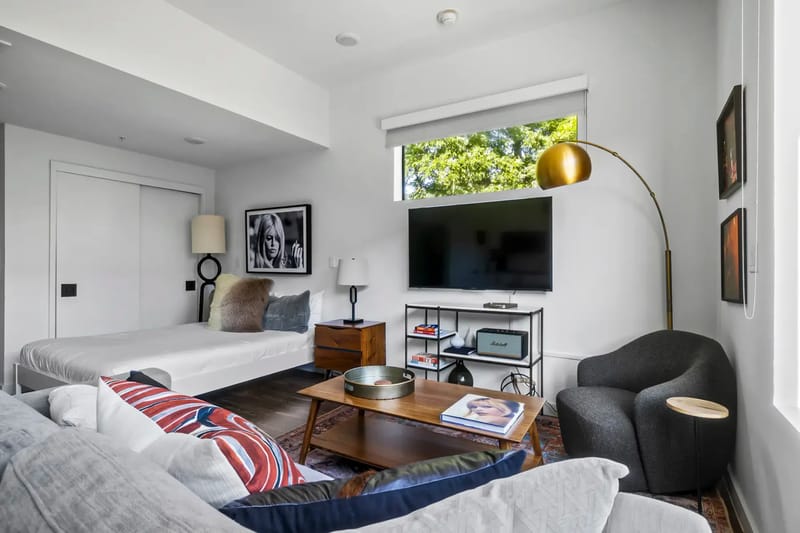 Sala de estar moderna con un sofá gris, un televisor montado en la pared, ventanas grandes, una mesa de centro de madera y obras de arte enmarcadas.