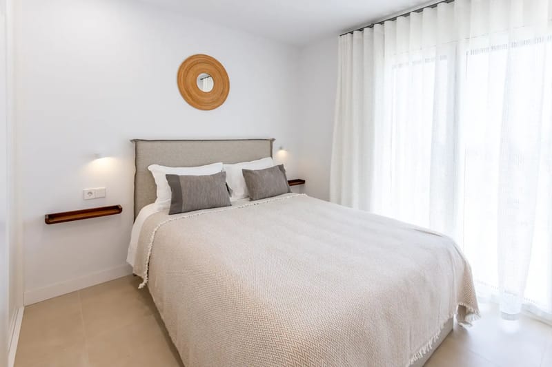 A neatly made bed with beige bedding, gray and white pillows, and sheer curtains.