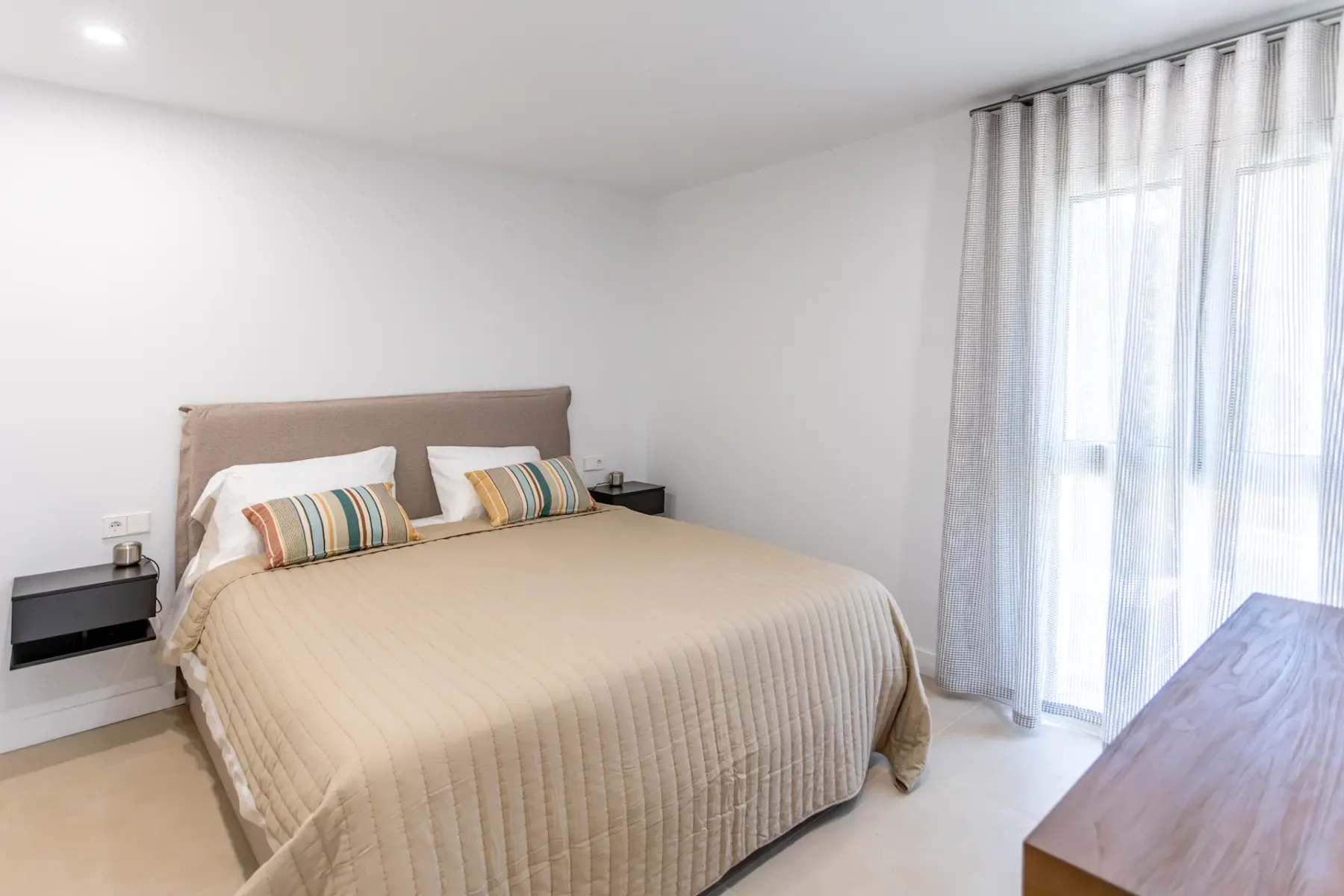A modern bedroom with a double bed, beige bedspread, striped pillows, and sheer curtains.