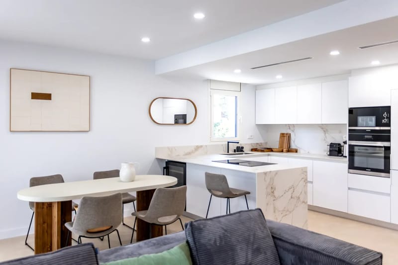Modern open-plan kitchen and dining area with white cabinets, marble countertops, a dining table with gray chairs, and minimalistic decor.