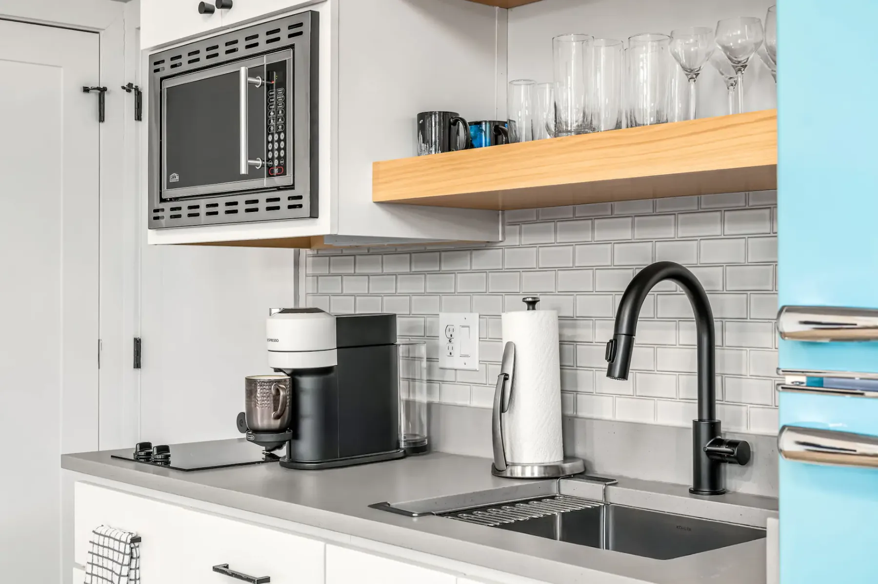 Rincón de cocina moderno con encimera gris, grifo negro, cafetera, microondas y estantes abiertos con vasos, sobre un fondo de azulejos.
