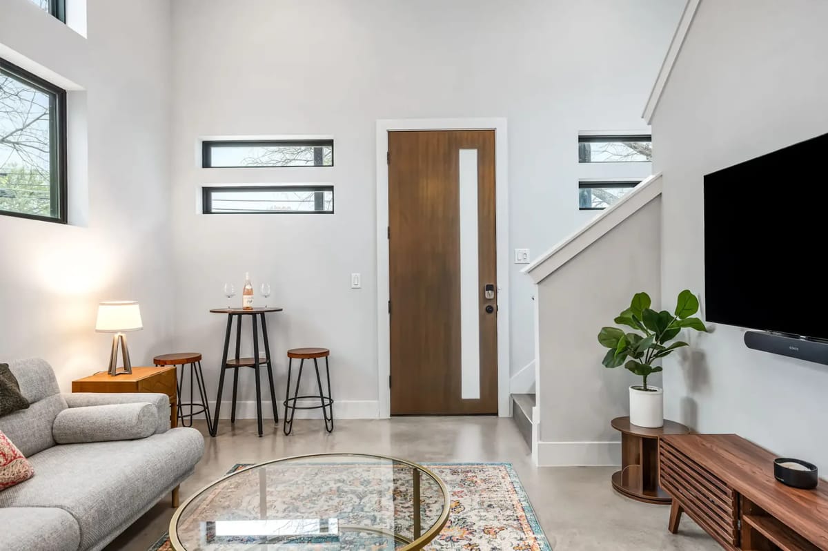 Modern living room with a sofa, glass coffee table, wall-mounted TV, potted plant, and two stools by a small table near the entrance.
