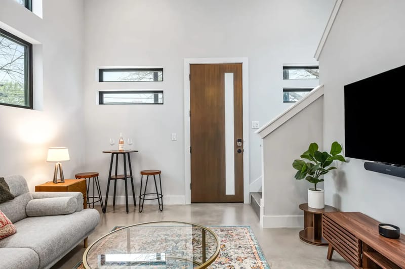 Sala de estar moderna con sofá, mesa de centro de vidrio, televisor montado en la pared, planta en maceta y dos taburetes junto a una mesa pequeña cerca de la entrada.