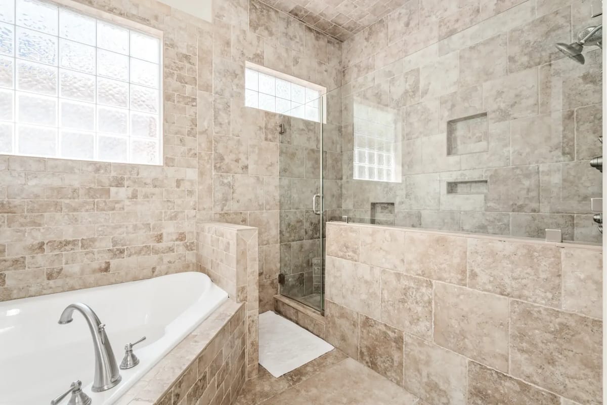 Baño moderno con una bañera de esquina, una ducha espaciosa con cerramiento de vidrio y paredes revestidas con azulejos de piedra clara.