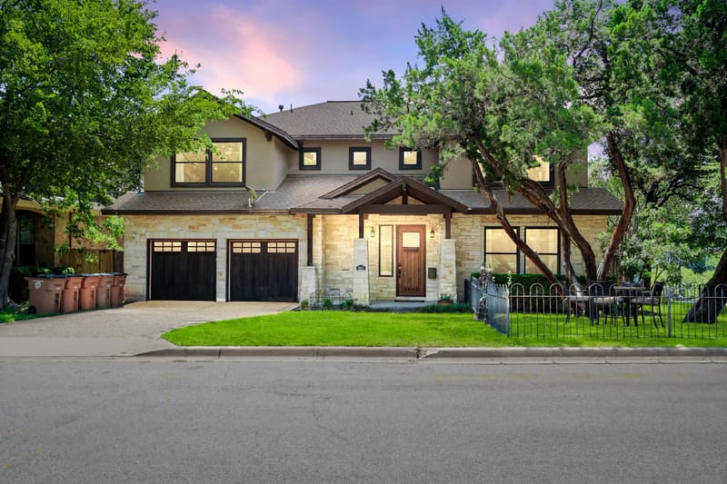 Two-story suburban house with a stone facade, double garage, and a small front yard, surrounded by trees, at sunset.