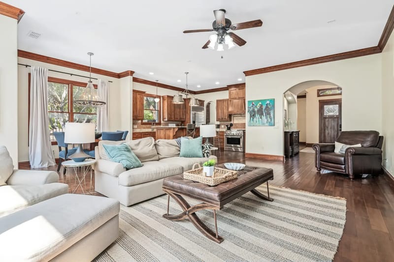 Spacious living room with hardwood floors, beige sofa, decorative fireplace, large windows, a ceiling fan, dining area, and natural light.