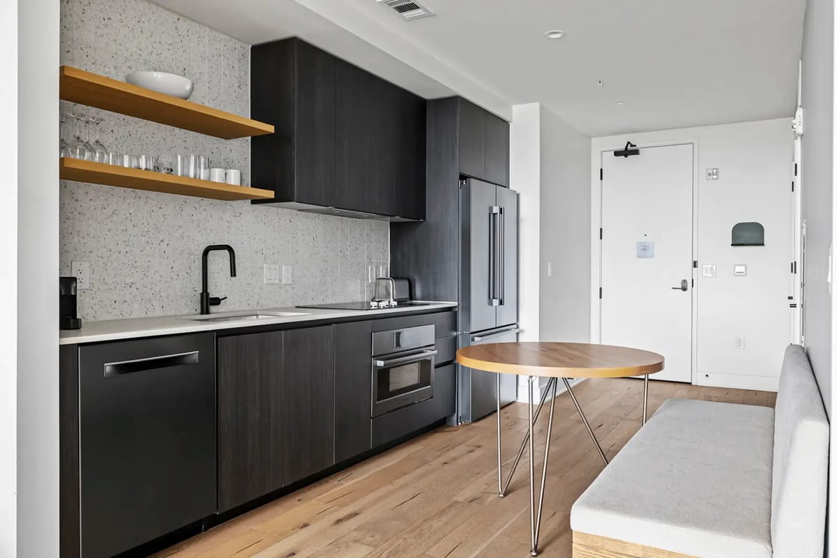 Cocina pequeña moderna con gabinetes oscuros, electrodomésticos empotrados, estantes abiertos y una mesa redonda de madera.