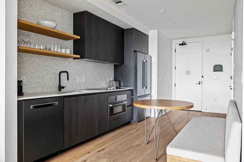 Cocina pequeña moderna con gabinetes oscuros, electrodomésticos empotrados, estantes abiertos y una mesa redonda de madera.