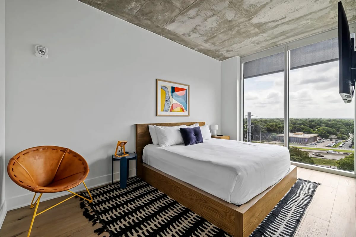 Dormitorio moderno con una ventana grande, cama de madera, ropa de cama blanca, alfombra estampada y arte colorido en la pared.