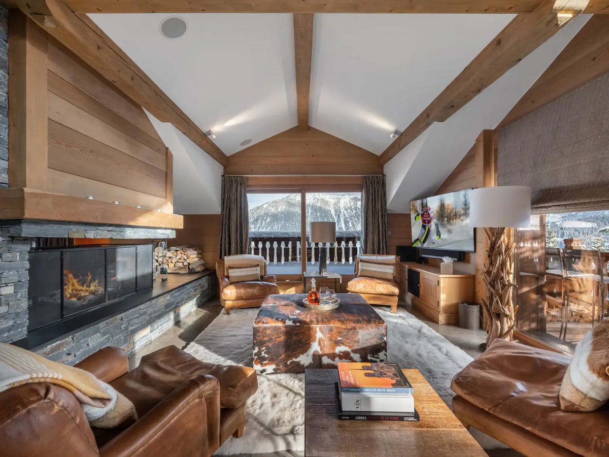 Acogedor salón con sillones de cuero, alfombra de piel de vaca, chimenea de piedra y grandes ventanales con vistas a las montañas nevadas.