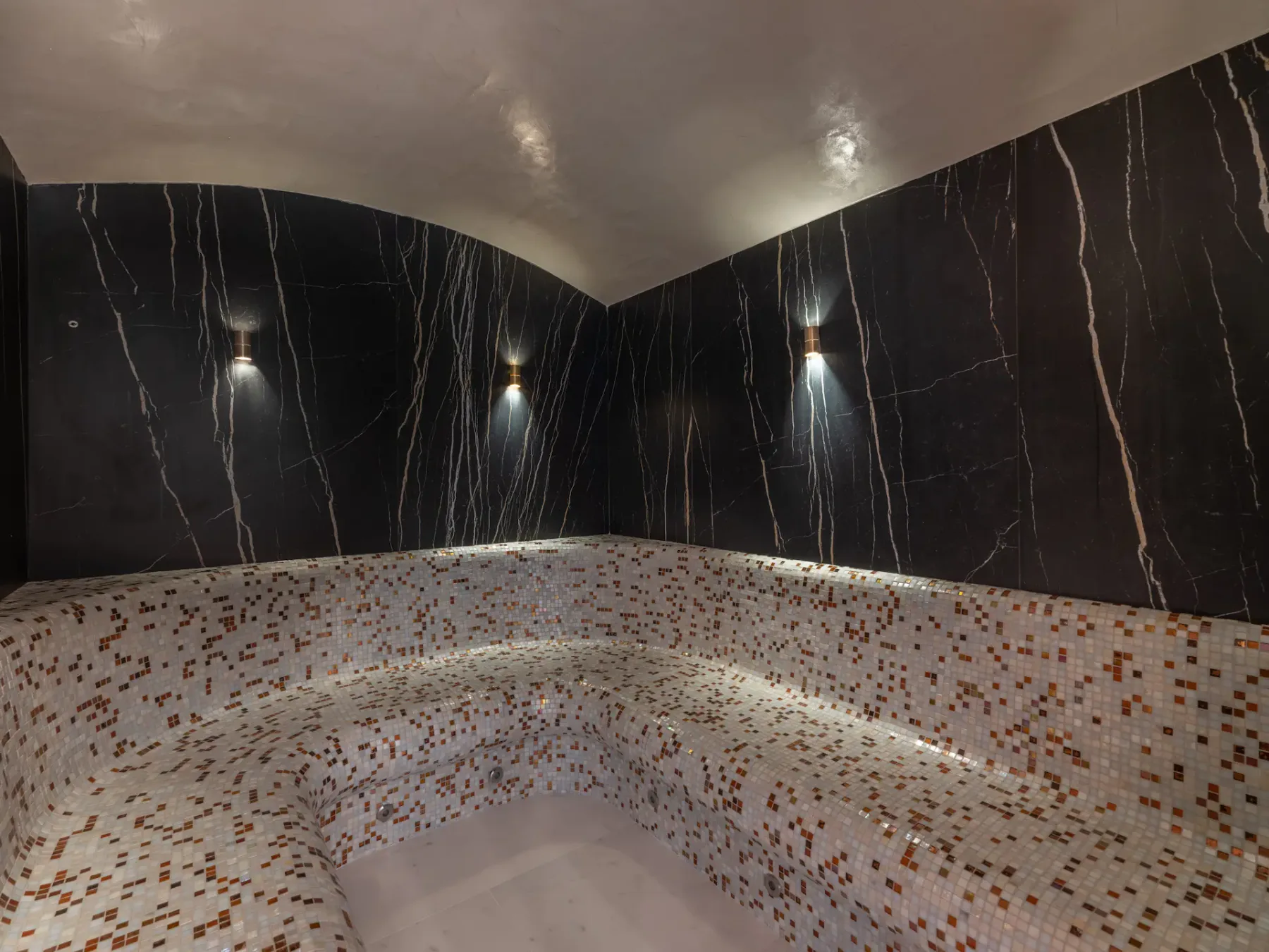 Baño de vapor con iluminación tenue, paredes de mármol oscuro y asientos revestidos de mosaico.