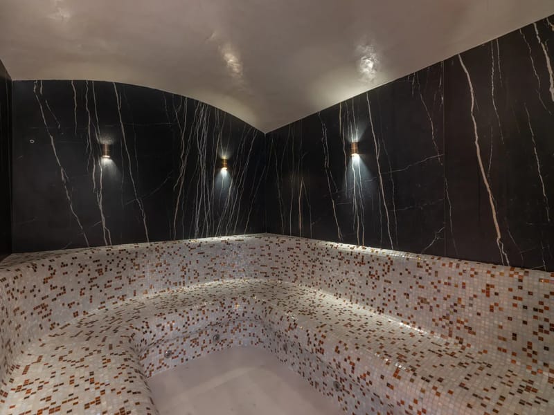 Dimly lit steam room with dark marble walls and mosaic tile seating.