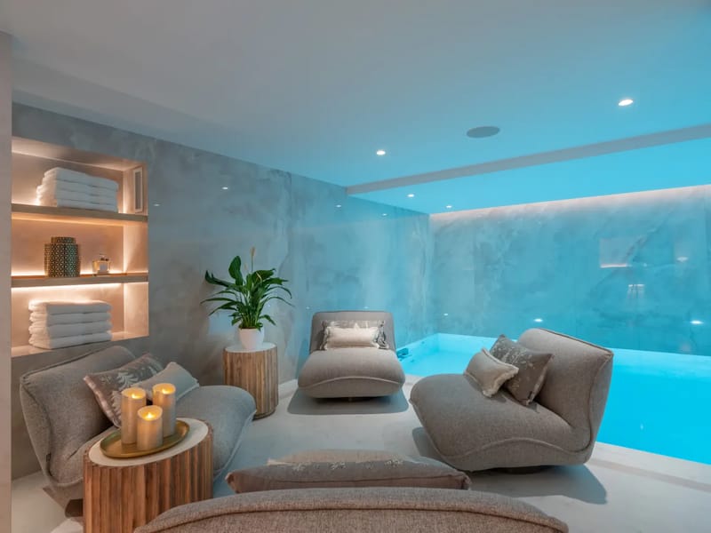 Luxurious spa room with a glowing indoor pool, plush lounge chairs, candles, and neatly stacked towels on shelves.