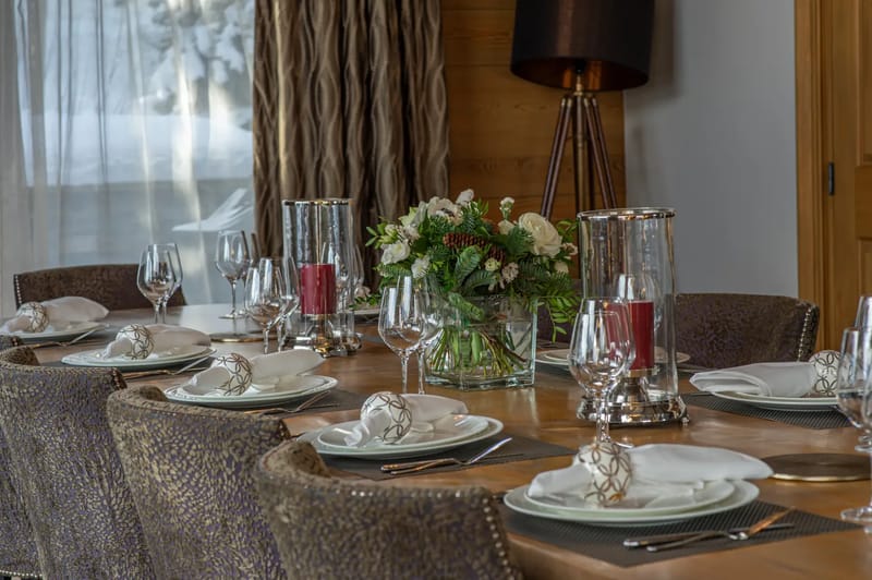Elegant dining table set for six with white plates, wine glasses, silver napkin rings, red candles, and a floral centerpiece.